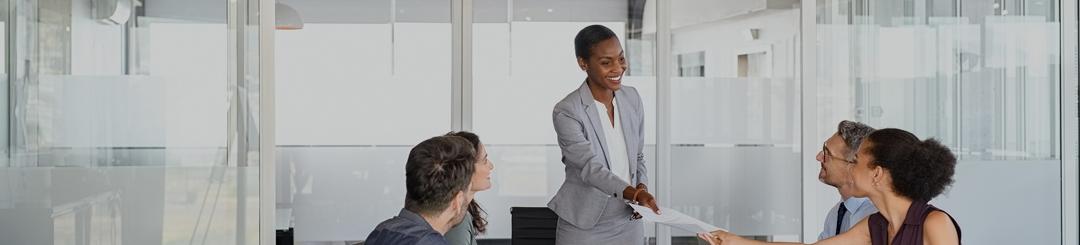 Woman handing paper to another woman sitting at conference table with a group of people.