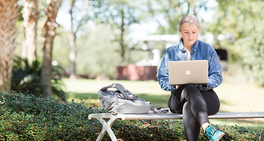 Student sitting outside on bench working on her laptop.